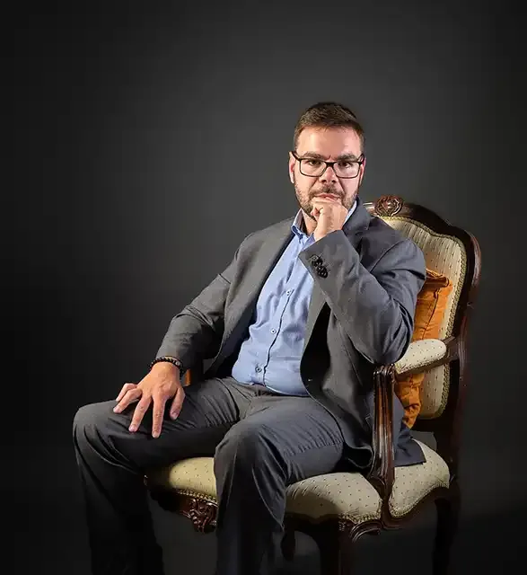 Marco Rodrigues sentado num cadeirão antigo sobre fundo escuro, de fato cinzento e camisa azul, a olhar para a câmara com a mão junto ao queixo.