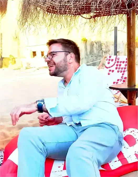 Marco Rodrigues sentado num puff vermelho sob um alpendre de canas, de camisa clara, a rir de lado.