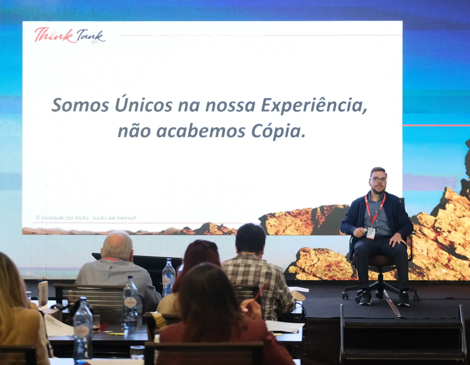 Marco Rodrigues sentado no palco de um evento Think Tank GPS, diante de uma plateia em mesas, com o diapositivo projetado atrás a dizer «Somos Únicos na nossa Experiência, não acabemos Cópia.»